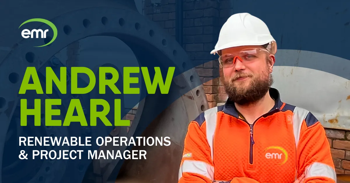 Andy, wearing a hard hat and safety vest stands in front of a wind turbine processing sign.
