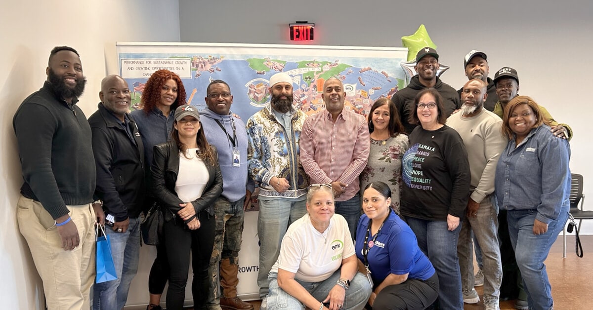 A group of sixteen people, standing and sitting in two rows, pose for a group photo indoors. They are smiling as part of the well-being event gathering.