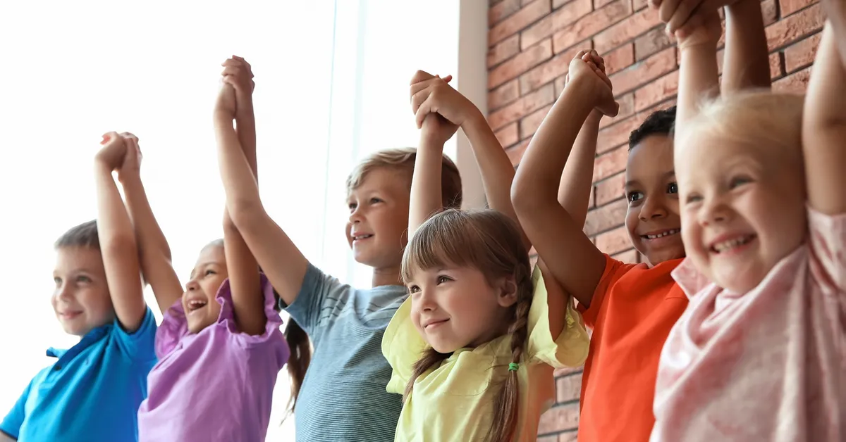 Een groep diverse kinderen die glimlachen en hun handen opsteken tegen een bakstenen muur.
