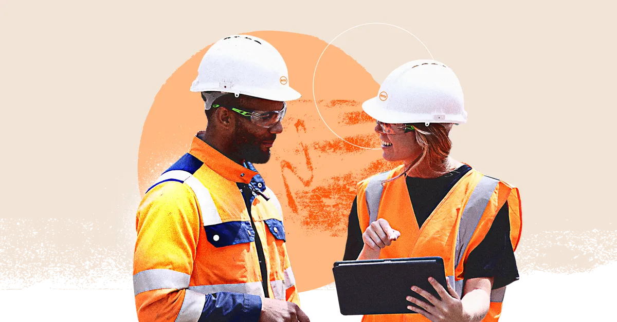 Two construction workers in hard hats and safety vests talking on a construction site.