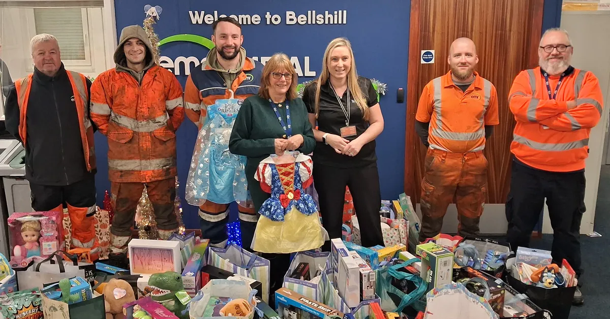 A group of people standing indoors in front of a blue wall with the text “Welcome to Bellshill” and part of a logo visible. Several individuals are wearing bright orange work uniforms, while two are dressed in colorful costumes resembling fairy-tale characters. In front of them, there is a large assortment of donated toys and gifts