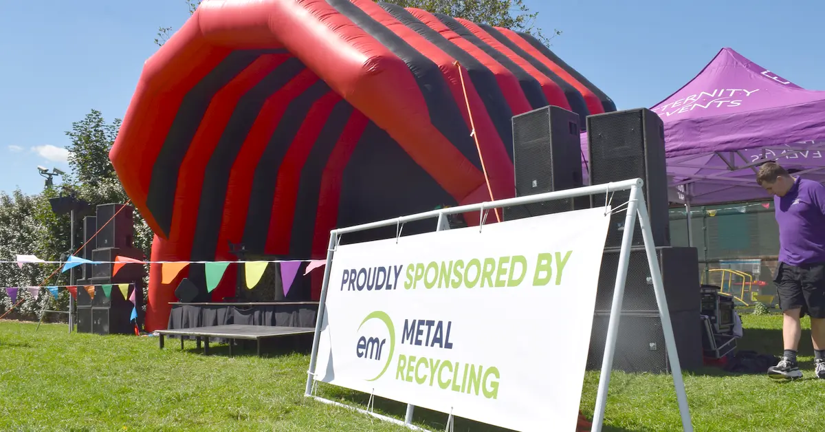 A vibrant red and black inflatable dome sits in a grassy outdoor. The dome has a segmented design with alternating black and red stripes. A white banner reading "PROUDLY SPONSORED BY EMR METAL RECYCLING" is attached to the side of the dome, featuring the EMR logo.