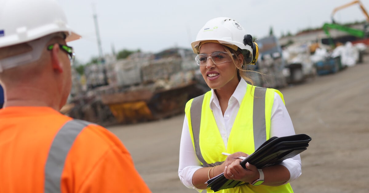 A woman wearing a white hard hat, protective glasses, and a neon yellow safety vest. She is holding a folder and is in conversation with a worker in an orange safety vest and white hard hat.