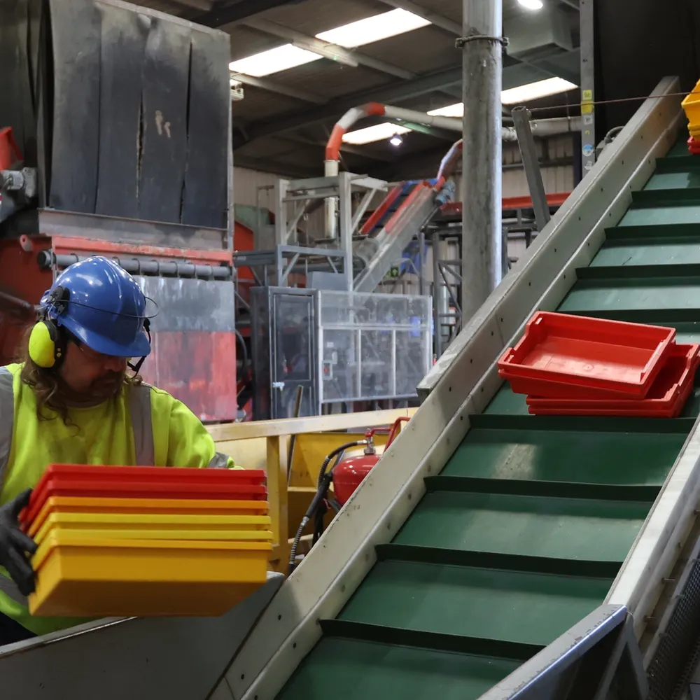 A person wearing a hard hat and safety vest stands next to a conveyor belt with red and yellow bins.