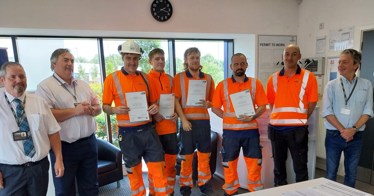 Group of EMR colleagues in orange safety gear receiving certificates, posing for a photo indoors with two officials in formal attire.