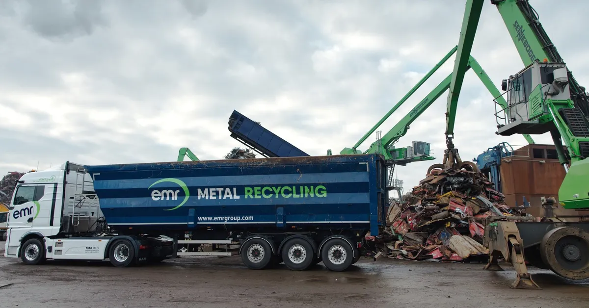 Een wit-blauwe vrachtwagen met "EMR Metal Recycling" op de zijkant staat geparkeerd naast een stapel schroot en een groene kraan onder een bewolkte lucht.