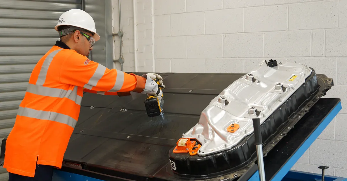 A worker in a high-visibility jacket and hard hat inspects an electric vehicle battery on a blue platform.