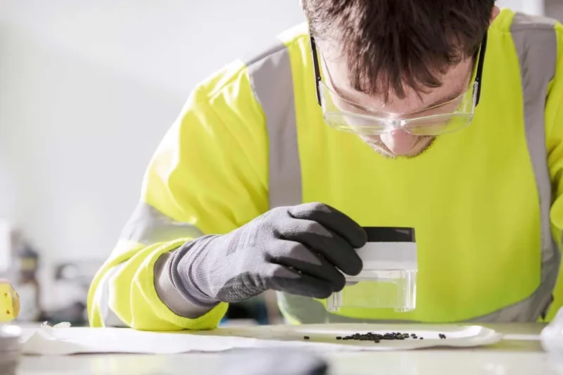 A person in a high-visibility jacket and safety goggles examining a petri dish with polymer pellets.