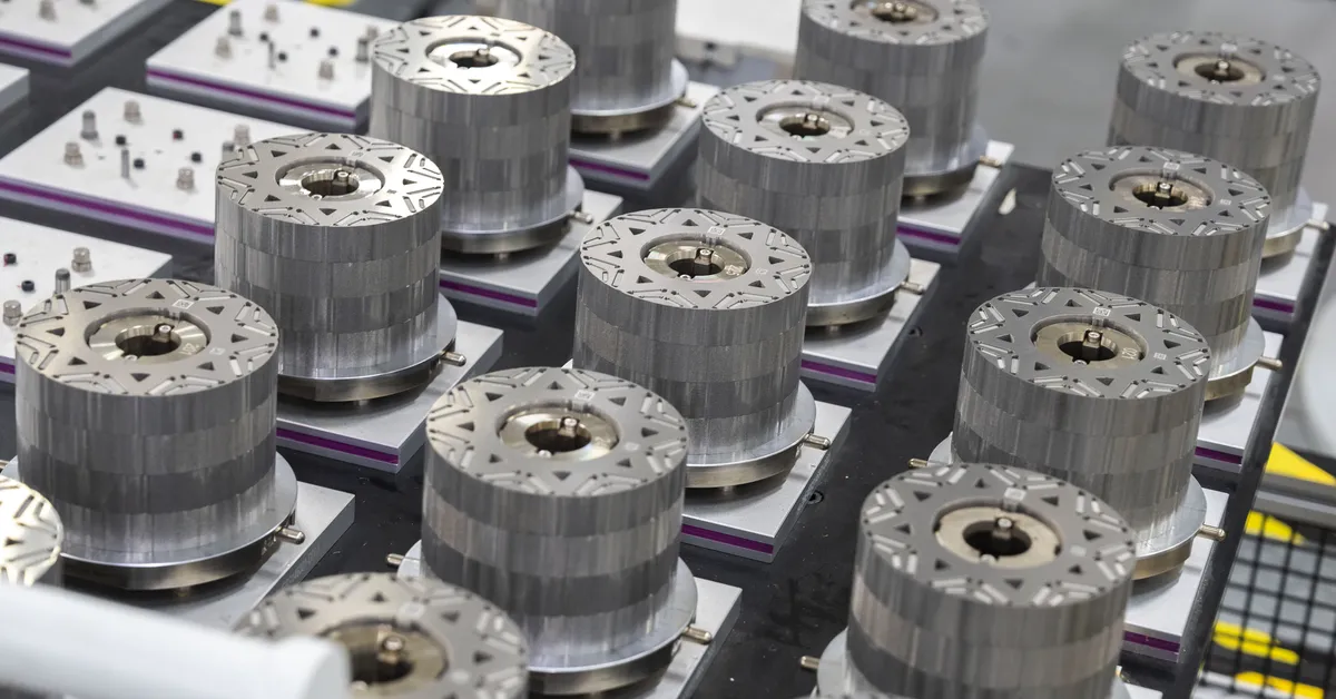 Rows of cylindrical metal components with patterned tops and central holes, each mounted on square bases, arranged on a surface in an industrial or manufacturing setting. Additional small parts are visible in the background.