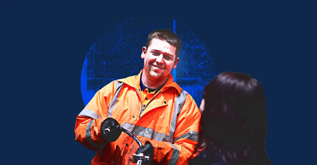 A person in an orange safety jacket holding a black tool and smiling at another person.