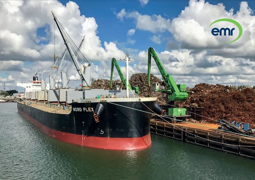 Een groot vrachtschip ligt aangemeerd in een haven. Het schip is zwart met een rode onderkant en is uitgerust met kranen aan boord. Op de kade ernaast verwerken twee grote groene kranen stapels schroot. De lucht is gedeeltelijk bewolkt met blauwe vlekken.