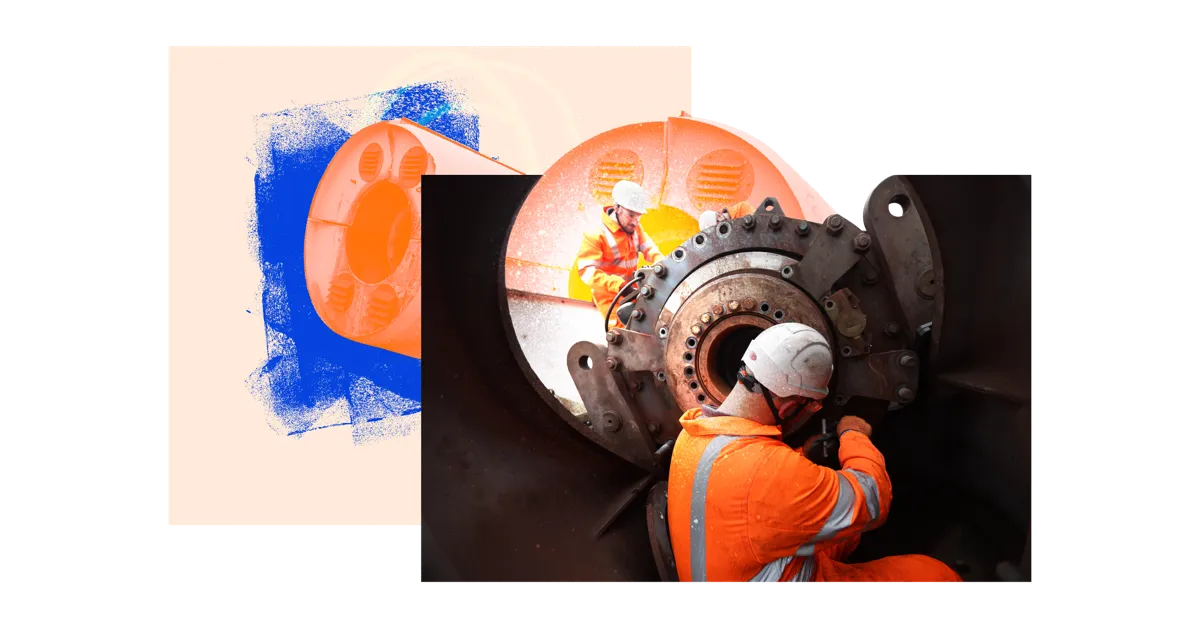 Two workers in hard hats and orange jumpsuits working on a large black machine.