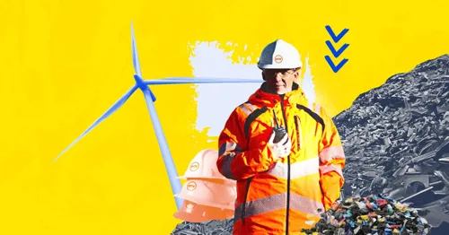 A person in a hard hat and safety jacket speaking at a podium in front of a wind turbine and a pile of scrap metal.