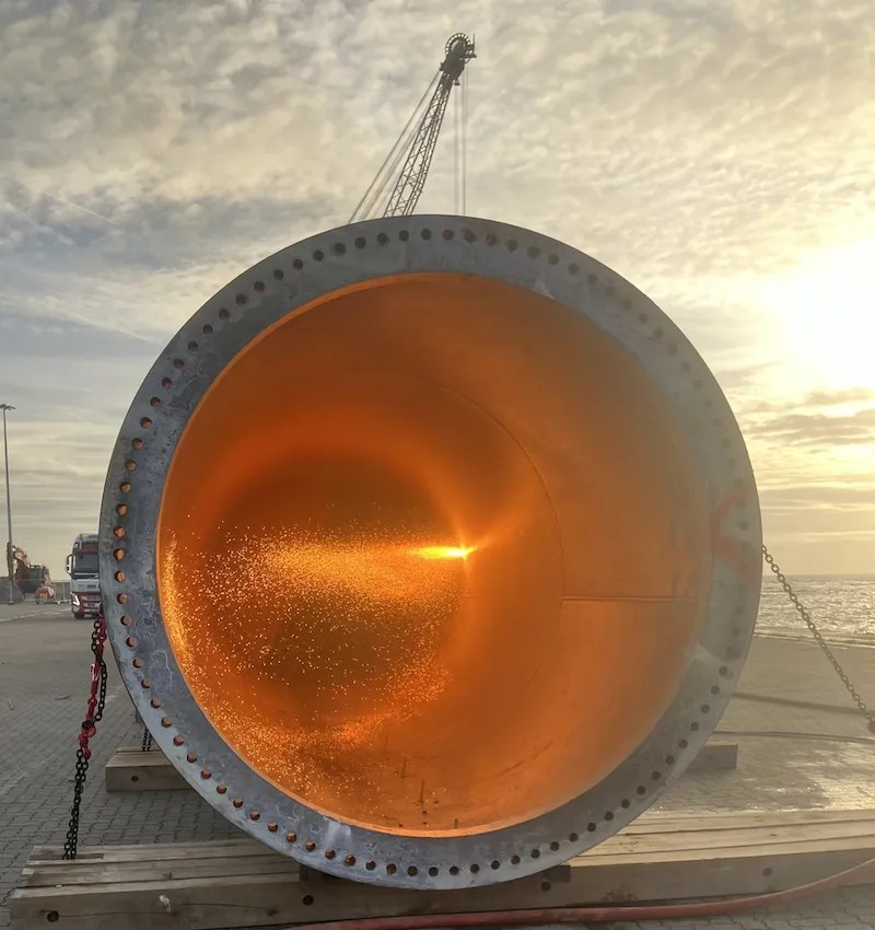 A crane lifting a large wind turbine cylinder near water.