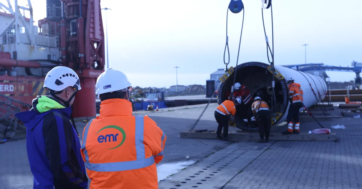 Two people in hard hats and safety vests stand near a large white wind turbine cylinder being lifted by a crane.